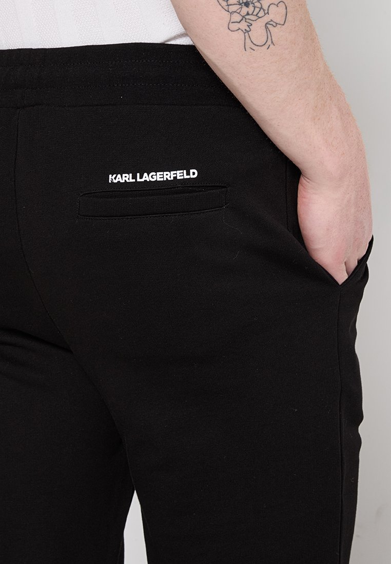 Person mit einem Unterarm-Tattoo, der schwarze Karl Lagerfeld-Hosen trägt, die Hand in der Seitentasche, die hintere Tasche sichtbar mit dem Markenlogo darüber.