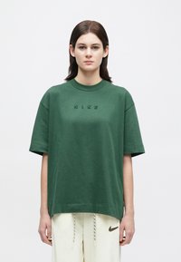 Άτομο που φοράει ένα πράσινο υπερμεγέθες μπλουζάκι Nike με κεντητό μαύρο λογότυπο και κρεμ φόρμες με λογότυπο Nike σε λεοπάρ σχέδιο.