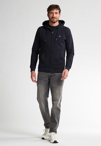 Marineblauwe zip-up hoodie met een capuchon, voorzakken en een trekkoord, gecombineerd met grijze jeans en witte sneakers. Eenvoudig en functioneel ontwerp.