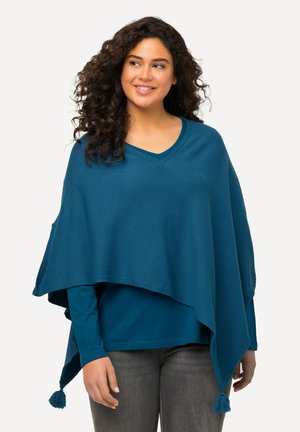 Türkiser Poncho mit V-Ausschnitt aus weichem Material. Verfügt über Fransendetails an den Ecken, übereinander getragen mit einem langärmeligen türkisen Oberteil.