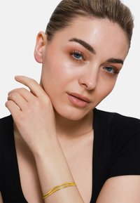 Bracciale dorato con due filamenti intrecciati, su uno sfondo chiaro. La mano del modello poggia sul collo, mettendo in mostra il bracciale.