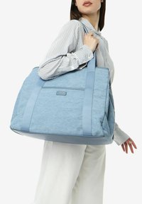 Sac fourre-tout bleu clair avec un corps en tissu texturé et des sangles lisses. Présente une étiquette frontale et un design spacieux pour une utilisation polyvalente.
