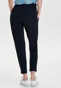 Pantaloni neri slim fit con una texture liscia, cintura elastica e tasche laterali, abbinati a sneaker bianche.