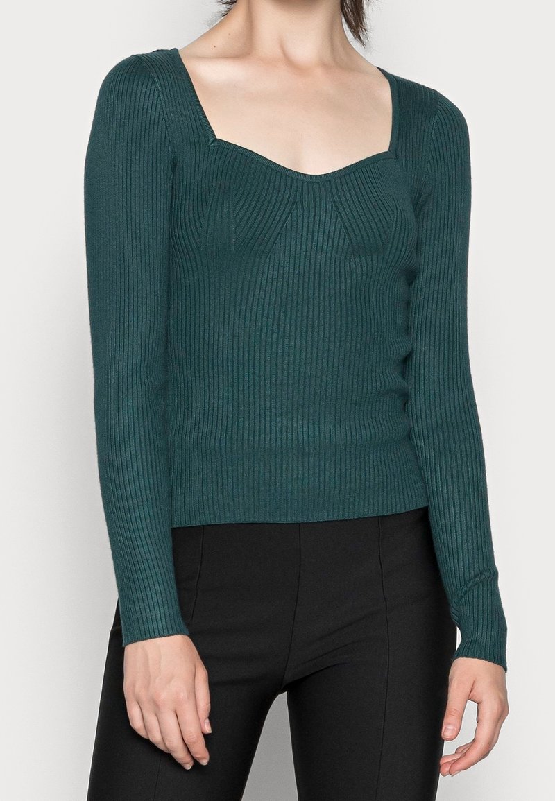 Femme portant un haut ajusté à manches longues côtelé bleu sarcelle avec un col carré et un pantalon noir, montrée du cou aux hanches.