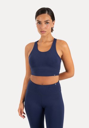 Frau, die ein marineblaues Sport-BH und passende hoch taillierte Leggings trägt, steht vor einem schlichten hellen Hintergrund.