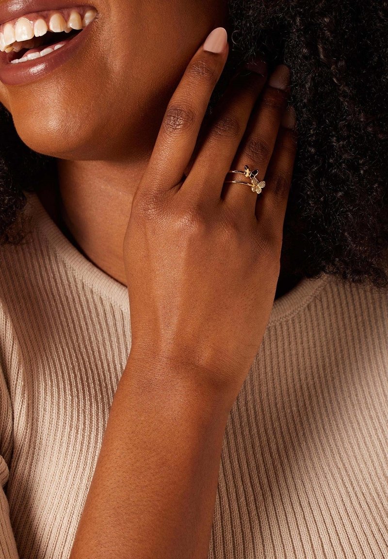 Gouden ringen met bloemaccenten liggen op een hand met een lichtbruine huidtint, met een gladde textuur en een lichtbeige ribbeltop.