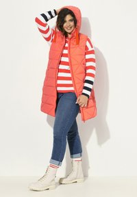 Gilet di corallo trapuntato con cappuccio, indossato sopra una maglietta a righe rossa e bianche, abbinato a jeans blu e stivali in pizzo color crema.