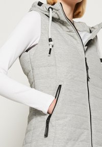 Light gray padded vest with a hood, featuring a zip front, adjustable drawstring, and two zippered side pockets. Ribbed white long-sleeve underneath.