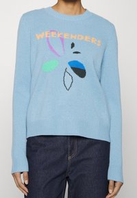 Pull en tricot bleu clair avec des poignets et un ourlet côtelés. "WEEKENDERS" en lettres orange. Présente des formes abstraites colorées : vert, bleu et noir.