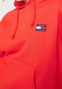 Felpa rossa in cotone con tasca frontale, cappuccio con lacci e un piccolo patch rettangolare del logo Tommy Jeans sul petto.