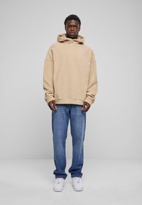 Beige hoodie met een halve ritssluiting, relaxte pasvorm en grote mouwen, gecombineerd met lichtblauwe rechte jeans en witte sneakers.