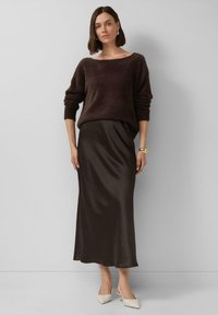 Ciemnobrązowy sweter dziewiarski z szerokim dekoltem, zestawiony z gładką, długą, ciemnobrązową satynową spódnicą. Modelka ma na sobie kremowe czółenka z ostrym noskiem.