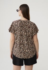 Blusa leopardata con maniche corte a volant, realizzata in tessuto leggero. Il retro presenta delle pieghe per un tocco di texture in più. Indossata con pantaloni corti neri.