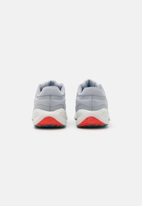 Zapatillas deportivas grises con un empeine liso, una media suela blanca y una suela exterior roja. Presentan sutiles líneas de diseño curvas y elementos de malla para mayor transpirabilidad.