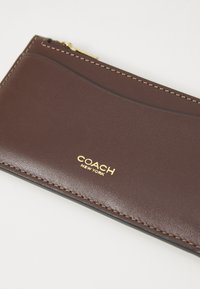 Braune Lederbörse mit glatter Textur, goldfarbenem Logo und kontrastierenden Nähten. Verfügt über einen Reißverschluss und ein geschwungenes Designelement.