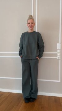 Persona ar gaišu bulciņu, tērpusies saskaņotā tumši zaļā džemperī un plašās biksēs, stāv un smaida pie bēša krāsas sienas.