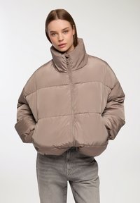 Giacca imbottita beige con colletto alto, dotata di una cerniera frontale, maniche larghe e polsini elasticizzati. Finitura liscia e lucida.