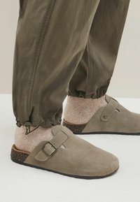 Sabots en suede gris avec un détail de boucle, présentant une semelle brune texturée, portés avec des chaussettes beiges à motifs et un pantalon vert olive ample.