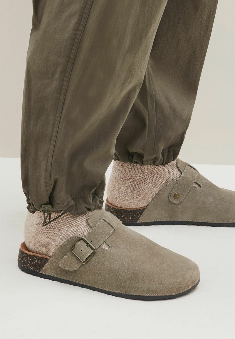 Sabots en suede gris avec un détail de boucle, présentant une semelle brune texturée, portés avec des chaussettes beiges à motifs et un pantalon vert olive ample.