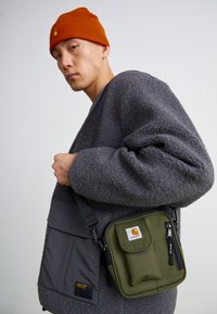Grå fleecejakke med oversized ærmer, parret med en khaki grøn crossbody taske med et logomærke og en lynlåslomme.
