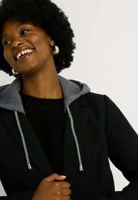 Blazer negro con sudadera gris, que cuenta con un cordón a rayas. La tela parece texturizada, con un bolsillo visible y un corte ajustado.