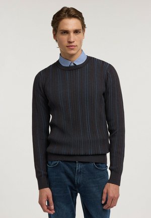 Jeune homme portant un pull rayé foncé par-dessus une chemise bleu clair à col et un jean bleu, debout devant un fond clair uni.