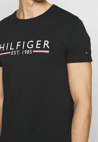 Camiseta de algodón negra con el texto blanco "HILFIGER EST. 1985", mangas cortas y un pequeño acento del logo tricolor en la manga.