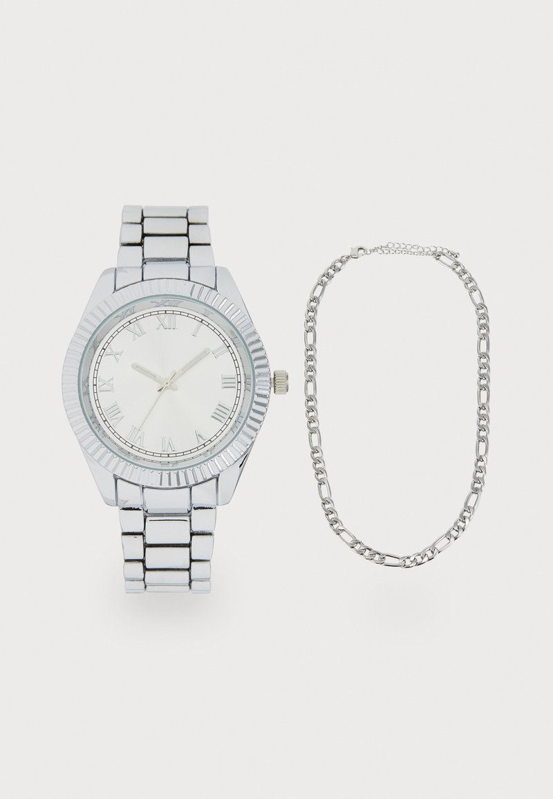 Montre-bracelet argentée avec un cadran rond, des chiffres romains, une lunette striée et un bracelet à maillons. Accompagnée d'un bracelet en chaîne argenté.