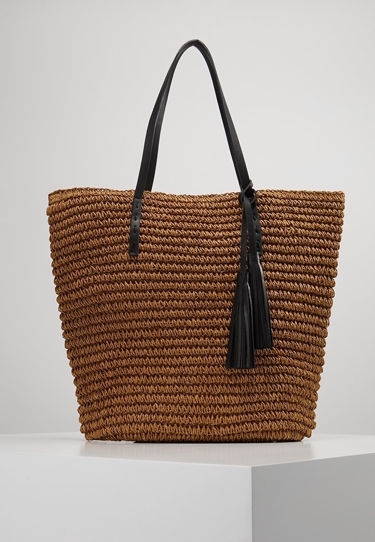 Tkana torba ze słomy w kolorze brązowym o konstrukcyjnej formie, z czarnymi skórzanymi rączkami i dekoracyjnym czarnym frędzlem.