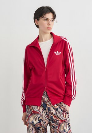 Person mit kurzen dunklen Haaren und Brille trägt eine rote Adidas-Trainingsjacke mit weißen Streifen und bunte Hosen mit Paisley-Muster.