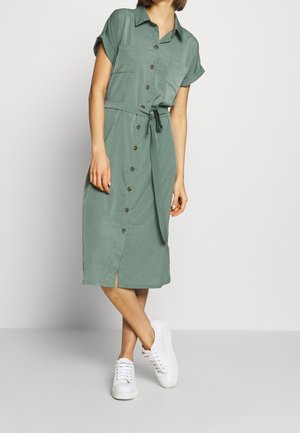 Robe chemise verte avec manches courtes retroussées, boutonnée à l'avant, deux poches poitrine, ceinture à la taille et fente latérale ; portée avec des baskets blanches.