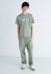 Joven de pie con las manos en los bolsillos, vistiendo una camiseta verde claro de Pepe Jeans, pantalones a juego y zapatillas blancas con acentos verdes.