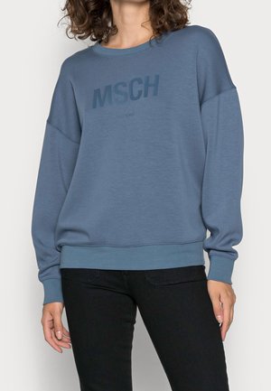 Femme portant un sweat bleu avec le texte "MSCH" sur la poitrine et un pantalon noir, debout devant un fond uni.