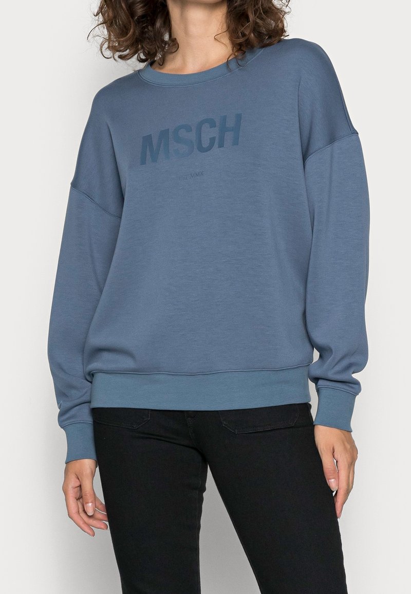 Femme portant un sweat bleu avec le texte "MSCH" sur la poitrine et un pantalon noir, debout devant un fond uni.
