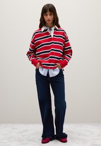 Rode en marineblauwe gestreepte sweatshirt met een witte kraag, gecombineerd met donkerblauwe flared jeans en roze instappers. Model staat met de handen in de zakken.