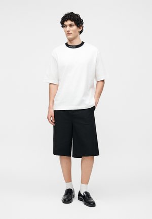 Junger Mann steht und trägt ein weißes Kurzarmhemd mit schwarzem Halsausschnitt, schwarze knielange Shorts, weiße Socken und schwarze Loafers.