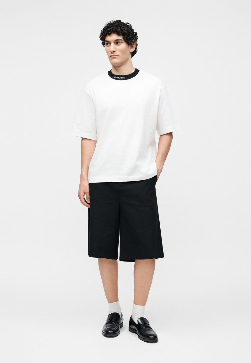 Νέος άνδρας στέκεται φορώντας λευκό κοντομάνικο πουκάμισο με μαύρο λαιμόκοψη, μαύρα σορτς μήκους μέχρι το γόνατο, λευκές κάλτσες και μαύρα παπούτσια τύπου loafers.