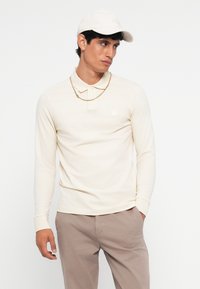 Lange mouwen roomkleurig poloshirt met een kraag en een logo-opdruk op de borst, gecombineerd met beige broeken en een bijpassende pet. Gouden kettingdetail zichtbaar.