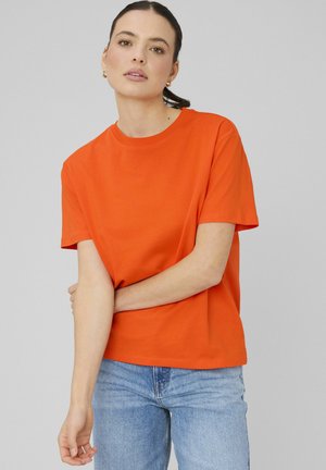 Femme aux cheveux foncés tirés en arrière, portant des boucles d’oreilles créoles dorées, un t-shirt orange à manches courtes et un jean bleu clair sur un fond gris uni.