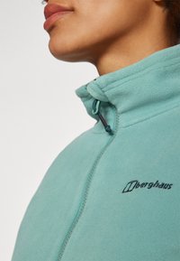 Veste en polaire bleu-vert clair avec un col haut, fermeture éclair et logo brodé noir sur la poitrine. Texture douce avec des accents de surpiqûre.