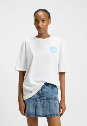 White cotton t-shirt with a blue sun graphic on the left chest, paired with a denim pleated skirt. Short sleeves and relaxed fit design.