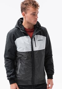 Giacca zip-up nera e grigia con cappuccio, caratterizzata da un design a blocchi di colore, tessuto testurizzato e una tasca con zip anteriore. Loghi stampati sul petto.
