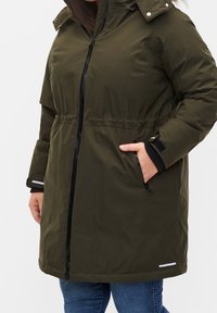 Parka impermeable de color verde oliva con capucha, cintura ajustable con cordón, cremallera negra y puños elásticos. Cuenta con dos bolsillos laterales.