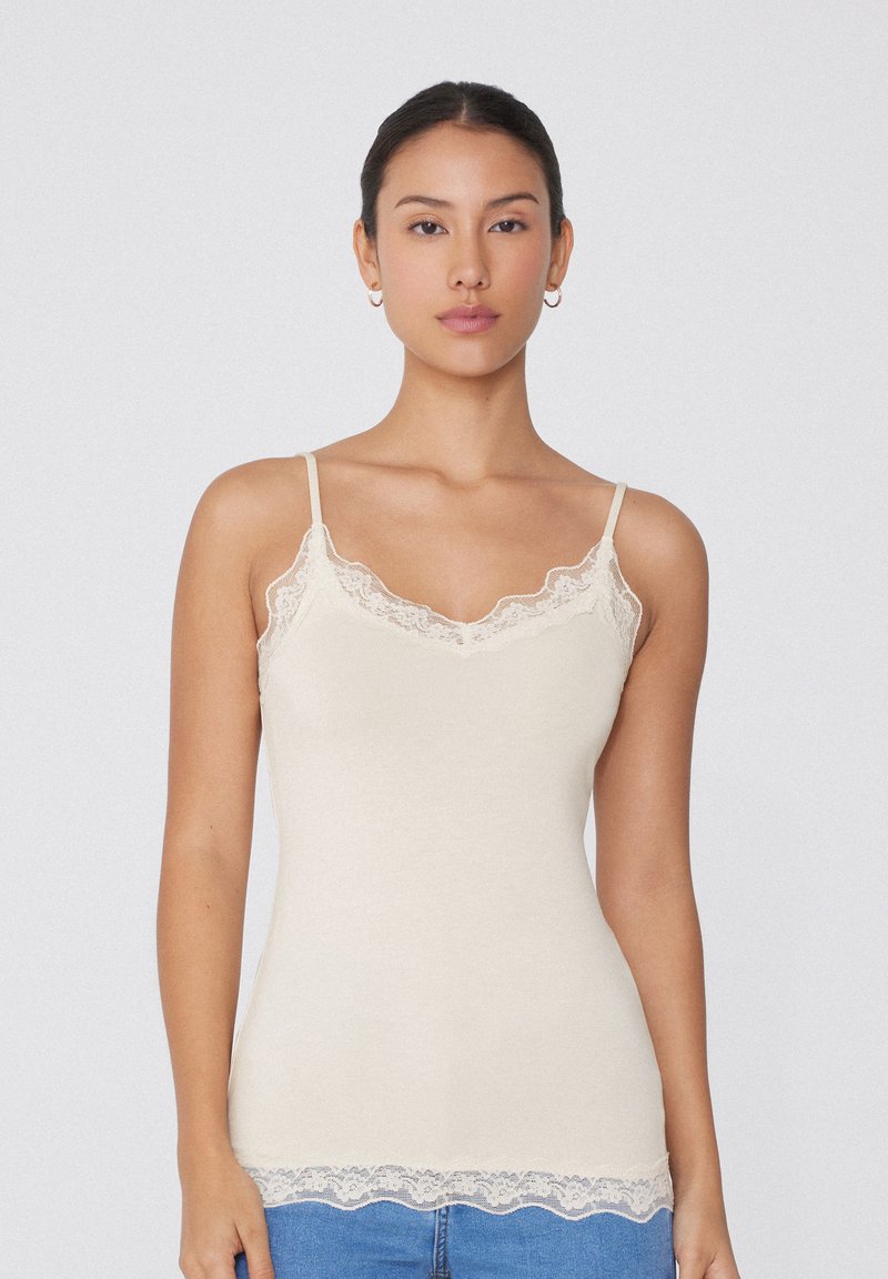 Beige Tops mit dünnen Trägern, Spitze entlang des Halsausschnitts und des unteren Saums, aus einem weichen, glatten Stoff gefertigt. Das Design ist figurbetont.