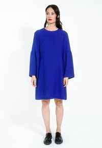 Rochie albastră, până la genunchi, cu mâneci lungi, evazate, decolteu rotund și țesătură fină. Purtați cu pantofi negri, tip slip-on.