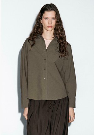 Camicia verde oliva, con bottoni, dal taglio rilassato, maniche lunghe e colletto a punta, abbinata a pantaloni scuri e larghi.