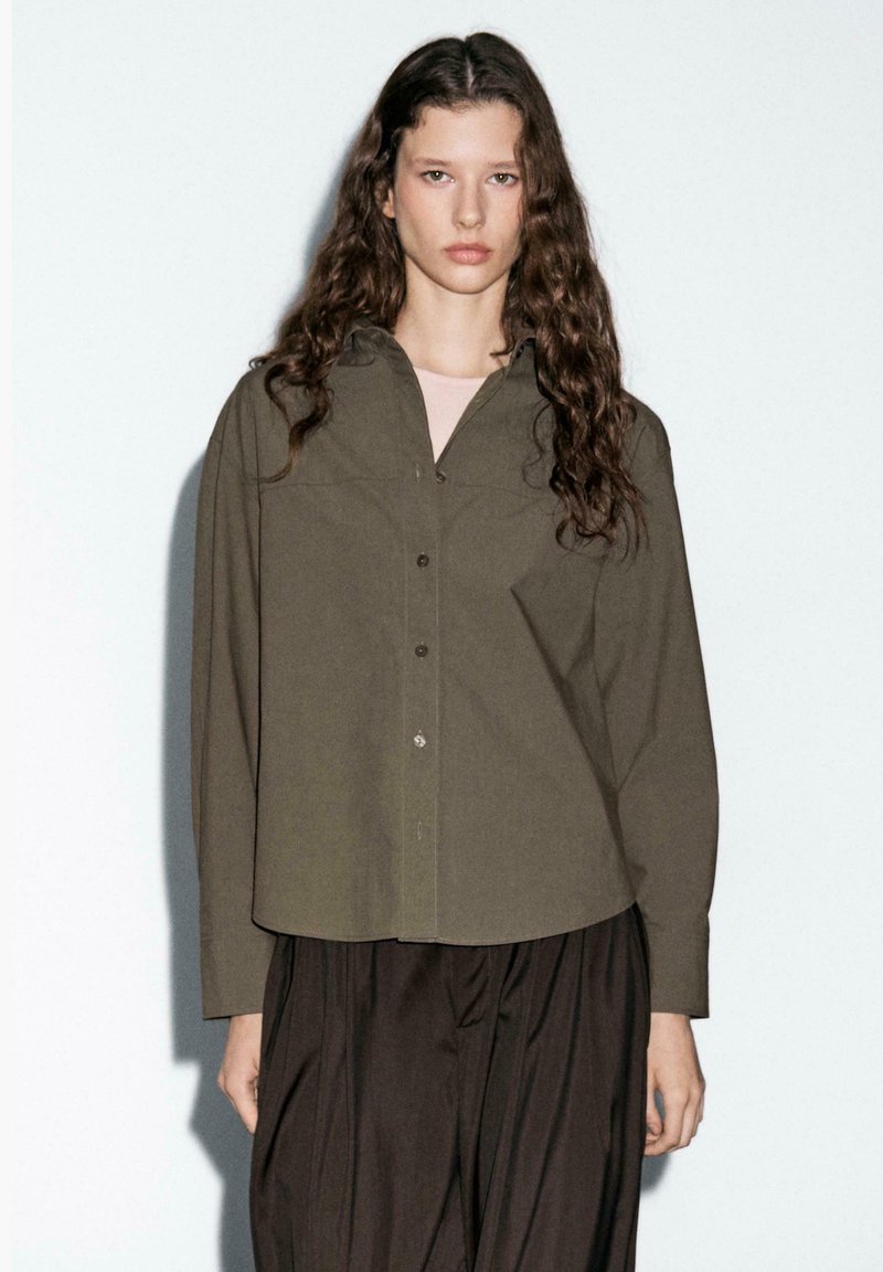 Olive green, button-up shirt with a relaxed fit, long sleeves, and pointed collar, paired with dark, loose-fitting pants.