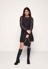 Robe noire avec de longues manches transparentes, motif à pois dorés, ourlet volanté, taille cintrée. Associée à un petit sac à main noir et des bottes jusqu'aux genoux.