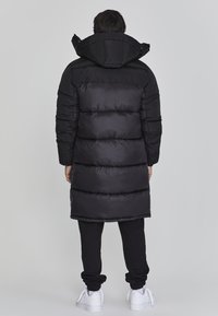 Schwarzer gepolsterter Mantel mit Kapuze, der horizontale Steppabschnitte und ein längeres Design aufweist. Getragen mit schwarzen Hosen und weißen Sneakers.