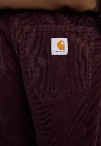 Bordeaux corduroy broek met een achterzak met een wit label en een oranje logo. De stof heeft een ribbel structuur en een gestructureerd ontwerp.